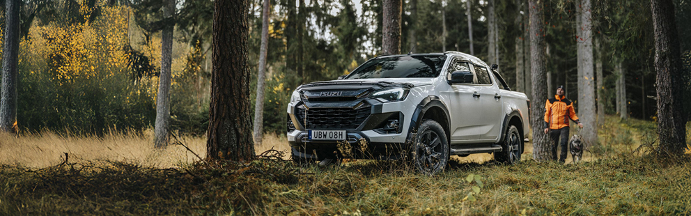 I en grönskande skog står en Isuzu D-Max redo medan en virkesplanerare och hund rör sig lugnt genom terrängen.