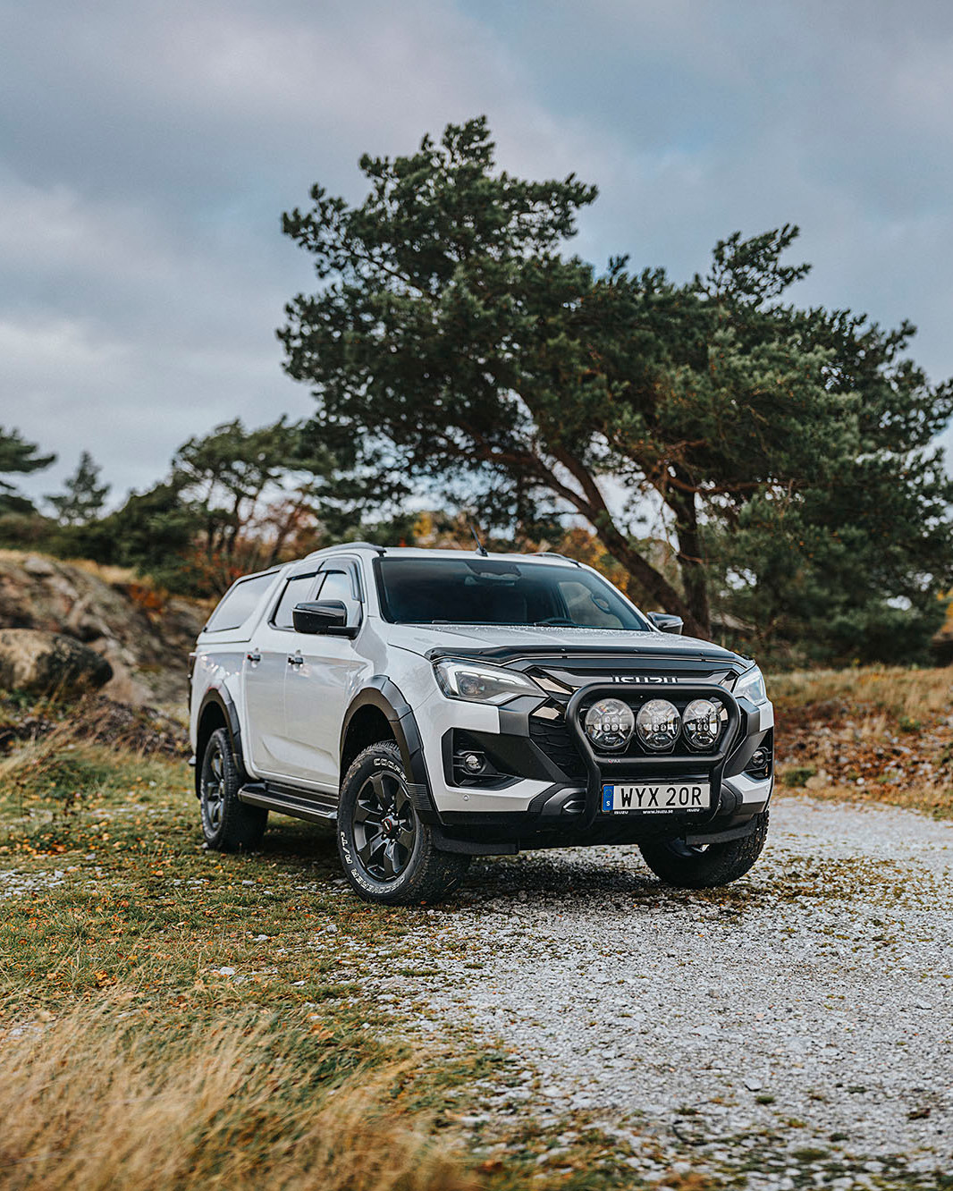 Vit Isuzu D-Max med flakkåpa ståendes på grusunderlag, med klippig natur i bakgrunden.