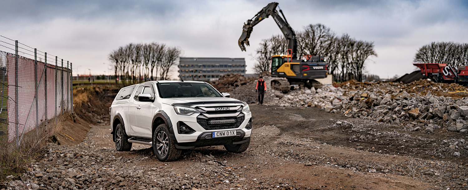 Isuzu D-Max Extended Cab parkerad på grus vid en rivningstomt, med grävmaskin i arbete i bakgrunden.