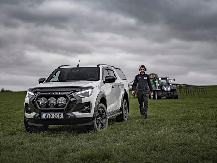 Vit Isuzu D-Max Double Cab med flakkåpa parkerad på lantlig åker – skördetröska i bakgrunden och lantbrukare går mot bilen.
