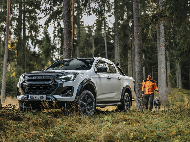 Isuzu D-Max står redo i skogen medan virkesinköparen och hunden rör sig längs stigen – skogligt arbete med rätt verktyg.
