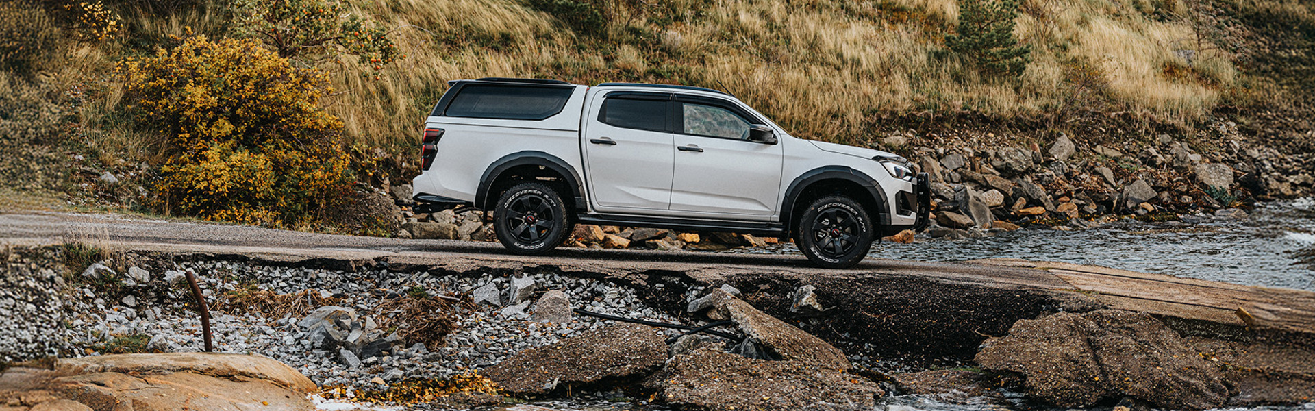 Isuzu D-Max kör ner för en ramp mot vattnet – en bro skymtar i bakgrunden, inramad av naturen.