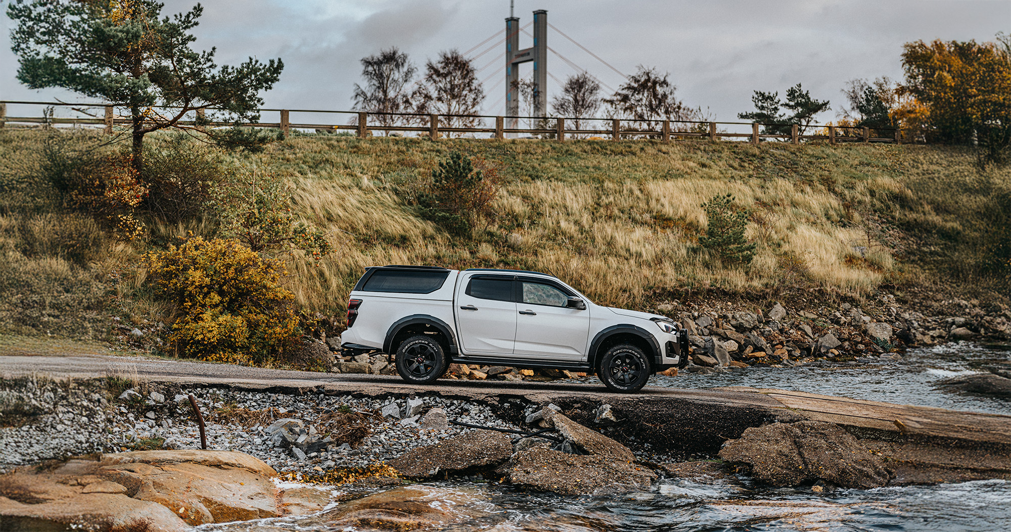 Isuzu D-Max kör ner för en ramp mot vattnet – en bro skymtar i bakgrunden, inramad av naturen.