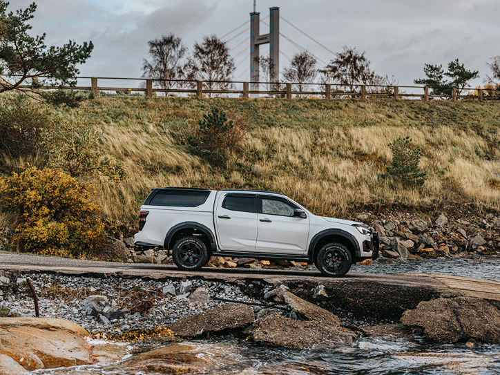Isuzu D-Max kör ner för en ramp mot vattnet – en bro skymtar i bakgrunden, inramad av naturen.