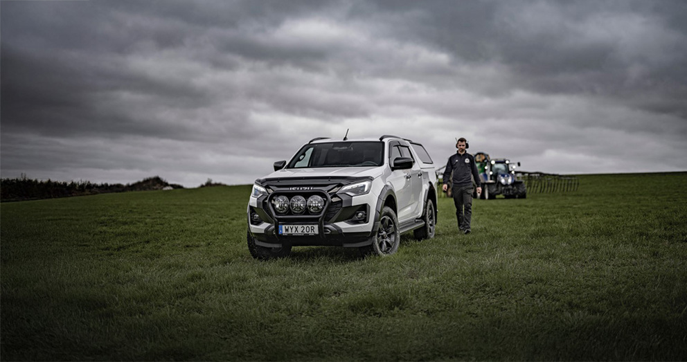 Isuzu D-Max i förgrunden på lantbruksmark, med skördetröska och bonde i arbete – ett fordon för den moderna lantbrukaren.