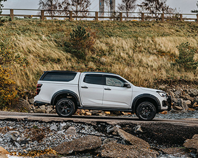 Isuzu D-Max på ramp mot en stilla sjö, med en bro i bakgrunden och omgiven av en dramatisk naturmiljö.