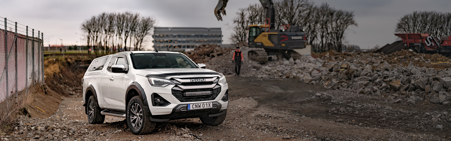 Isuzu D-Max Extended Cab parkerad på grus vid en rivningstomt, med grävmaskin i arbete i bakgrunden.