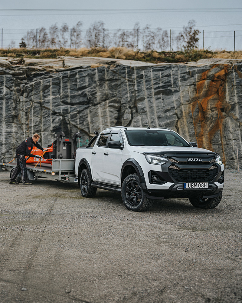Vit Isuzu D-Max utrustad med rolltop och flakbåge, drar ett släp med 3,5 ton last – en hydraulhammare på flaket.