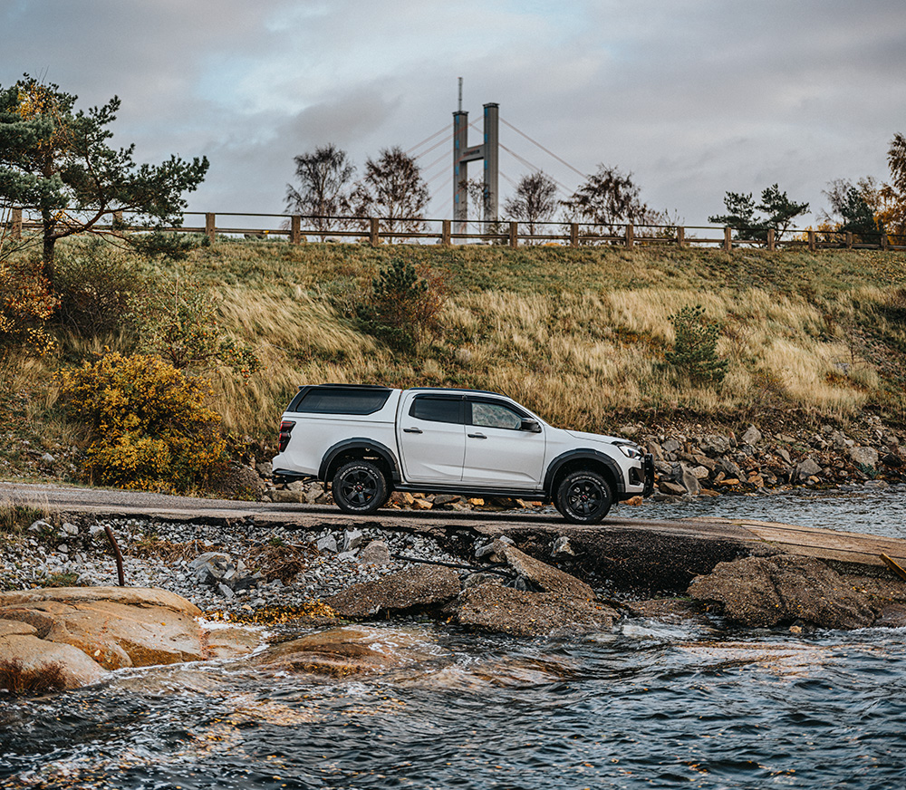 Isuzu D-Max kör ner för en ramp mot vattnet – en bro skymtar i bakgrunden, inramad av naturen.
