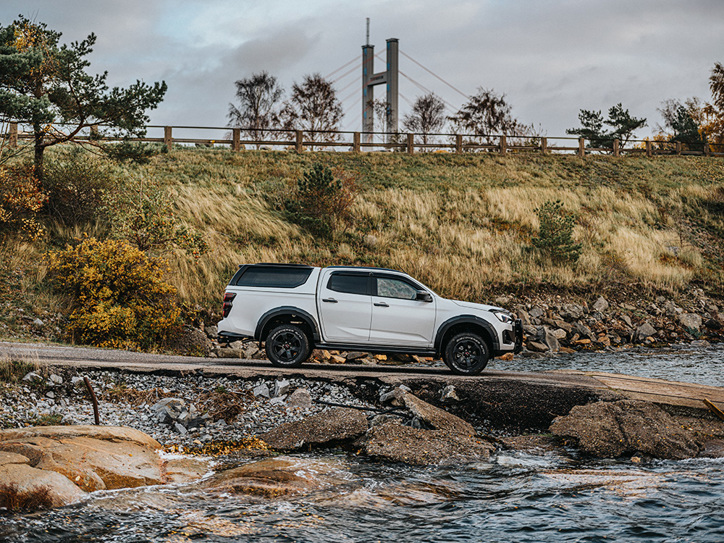 Isuzu D-Max kör ner för en ramp mot vattnet – en bro skymtar i bakgrunden, inramad av naturen.