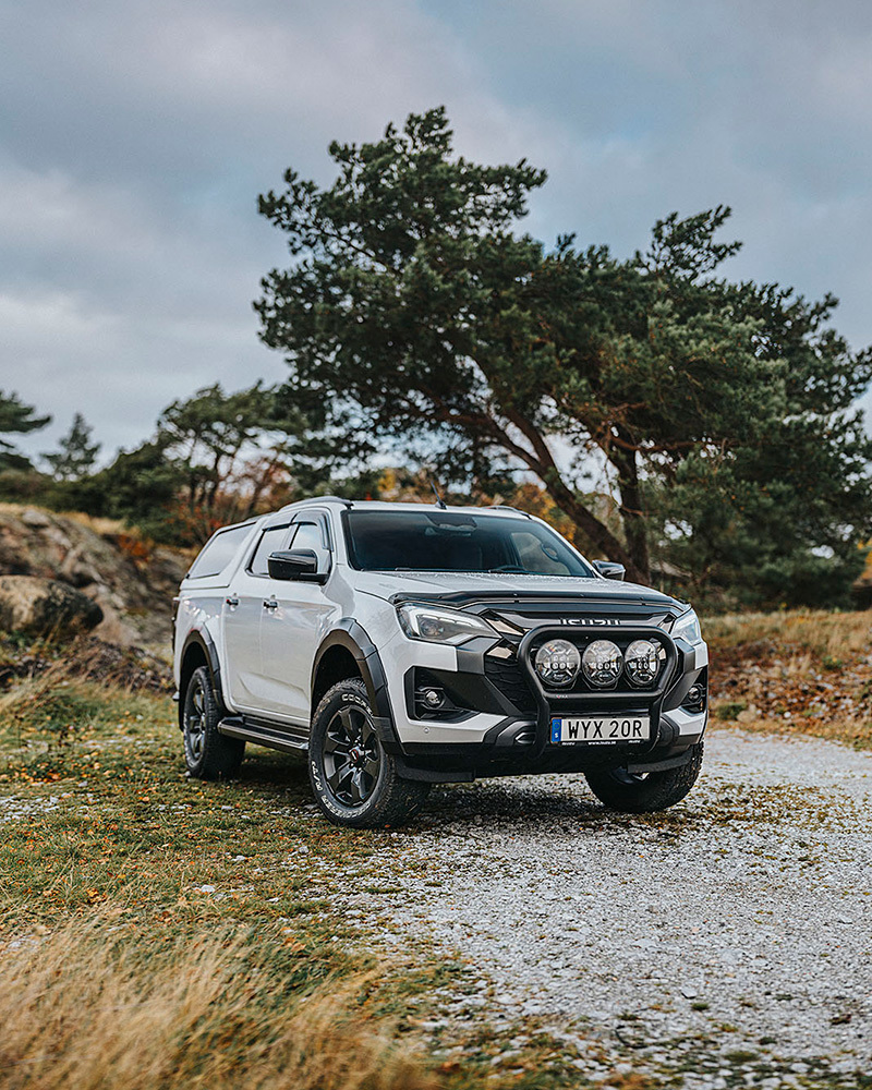 Vit Isuzu D-Max med flakkåpa ståendes på grusunderlag, med klippig natur i bakgrunden.